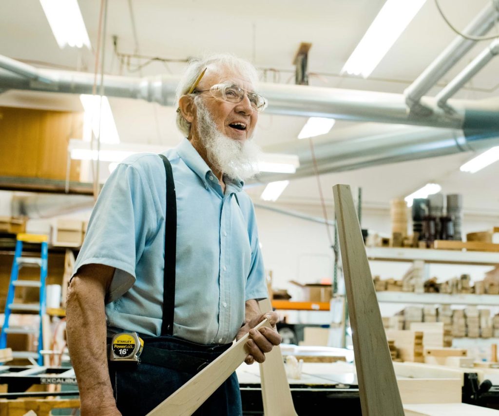 An elderly Amish craftsman with a white beard and suspenders holding a wooden piece in a workshop