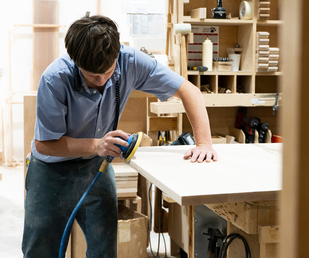 A craftsman sanding a wooden board with a power sander in a woodworking shop